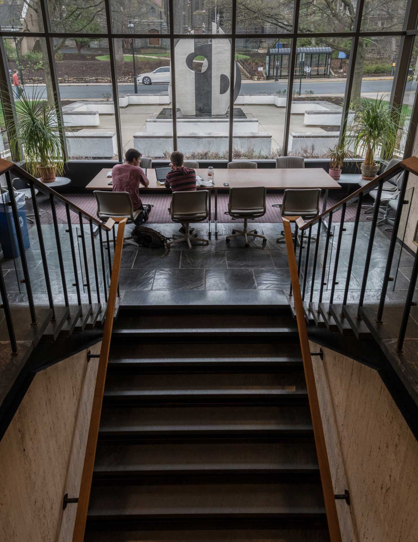 Two men are sitting ant talking at a shared desk in front of the central staircase.