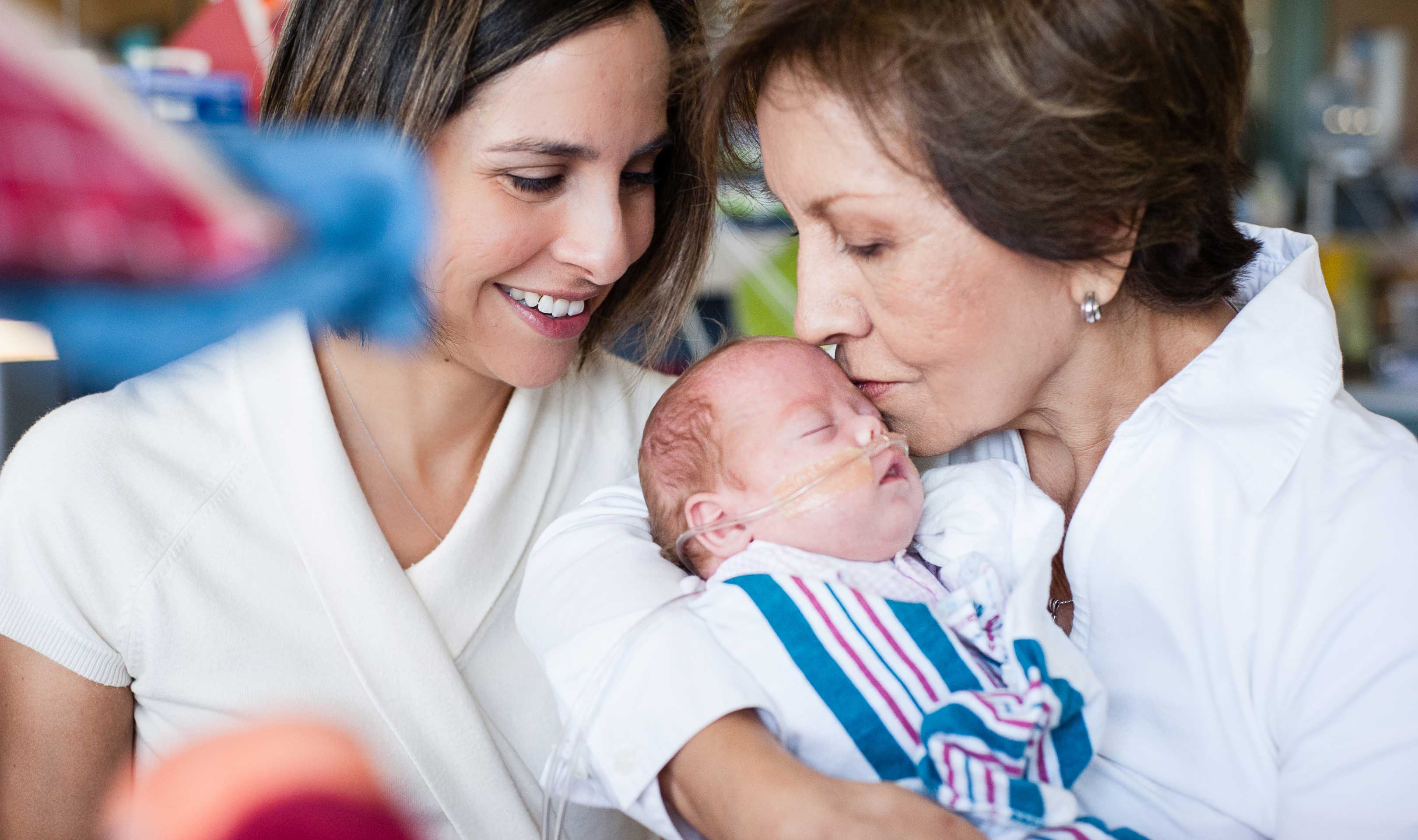 A grandmother is holding a sleeping baby while another woman looks on, smiling at the baby.