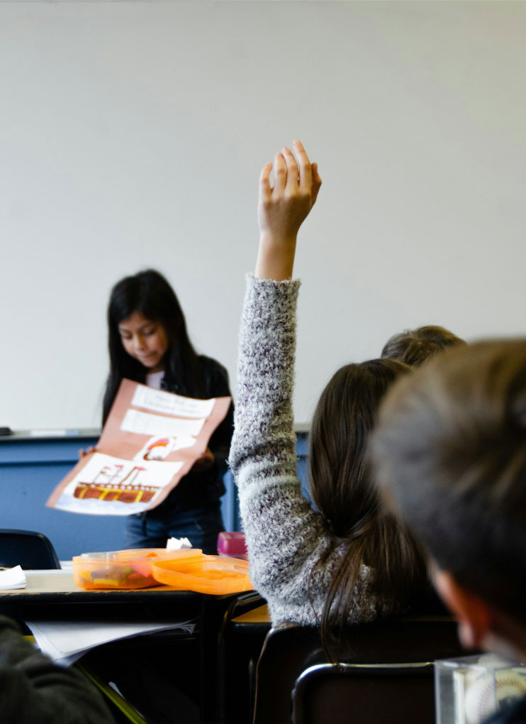 A kid is raising her hand while other kid is presenting in a classroom