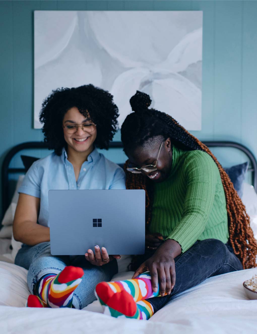 Two women are sitting on the bed, looking at a laptop and laughing.