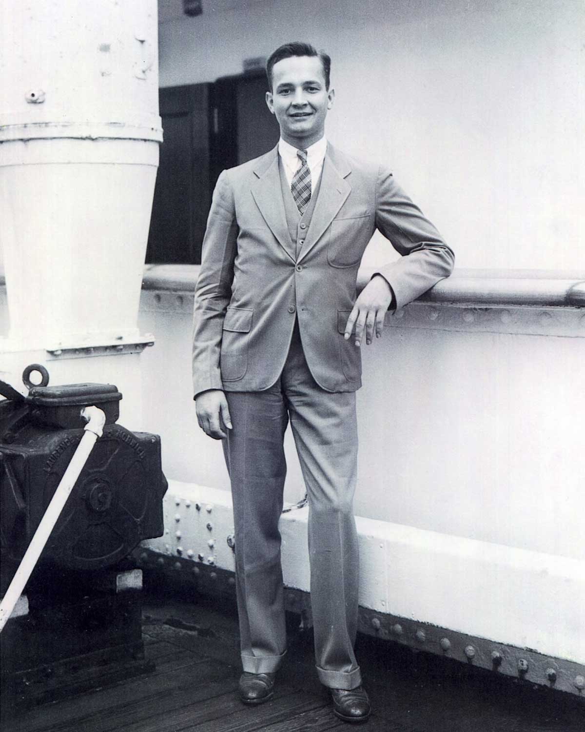 A young man in a suit is leaning his arms on the railing of a ship, smiling.