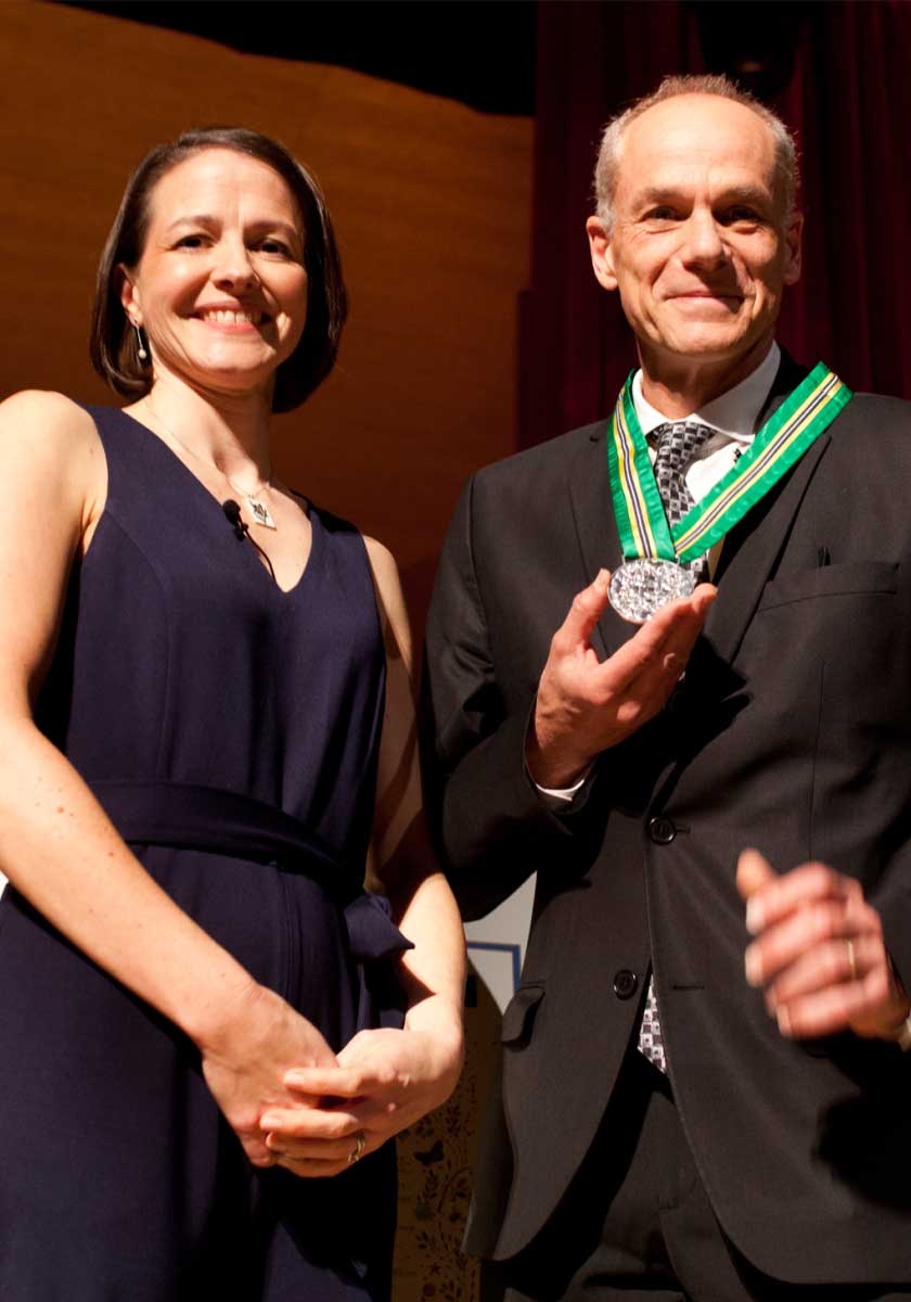 A middle-aged woman and man are smiling while looking ahead, and the man is holding a silver medal hanging around his neck.