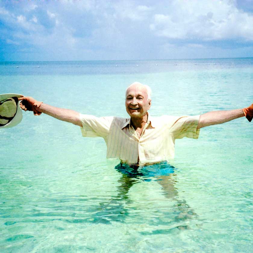 An elderly man with white hair is in the water, raising his arms in joy and laughing.